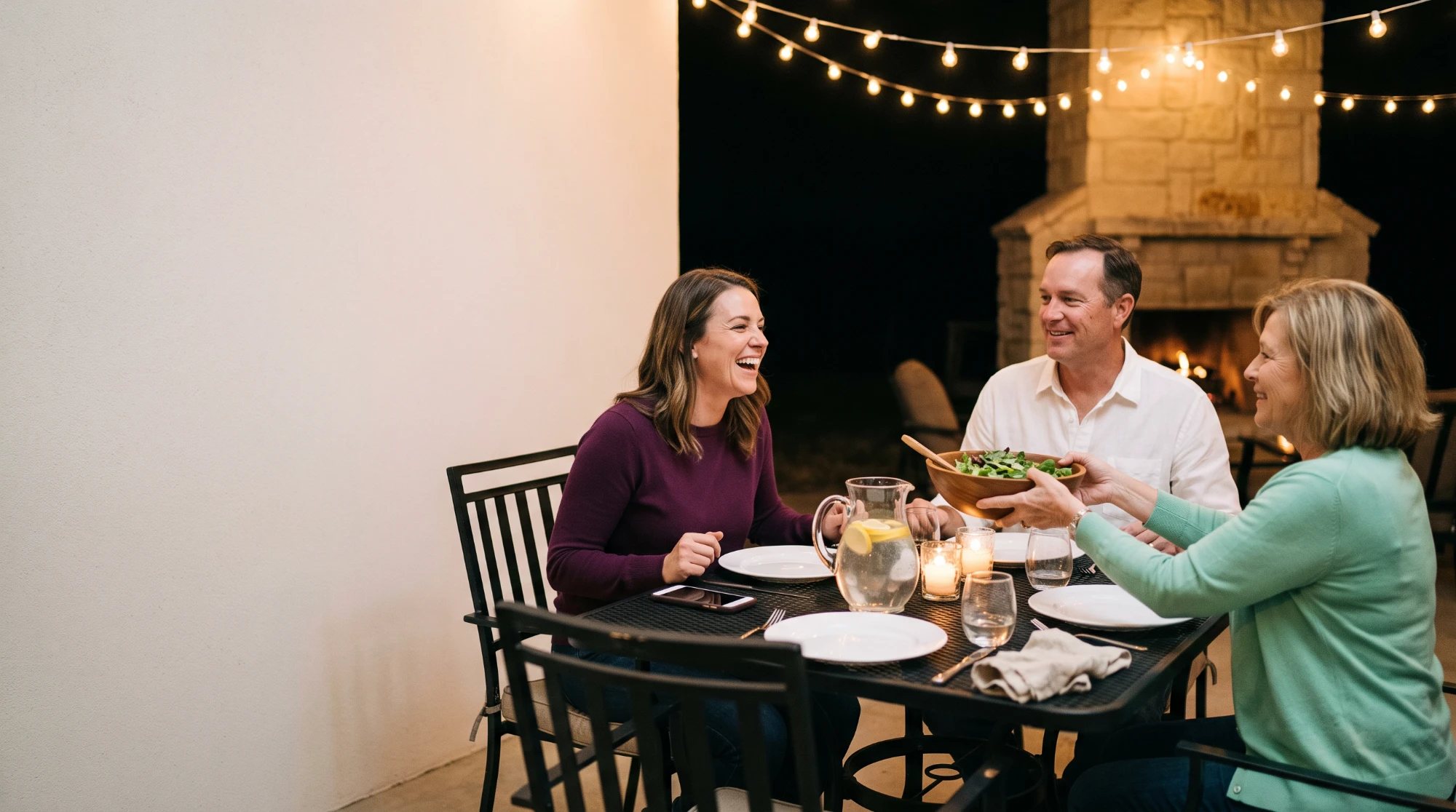 A woman in a plum sweater laughing at dinner with a man in a white shirt and an older woman in a mint cardigan who passes a salad bowl, at a black metal table on a night patio under string lights with a stone fireplace behind them and a phone beside her plate